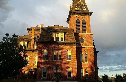 Large brown brick building with many arched windows and tall clock tower against a grey cloudy sky.