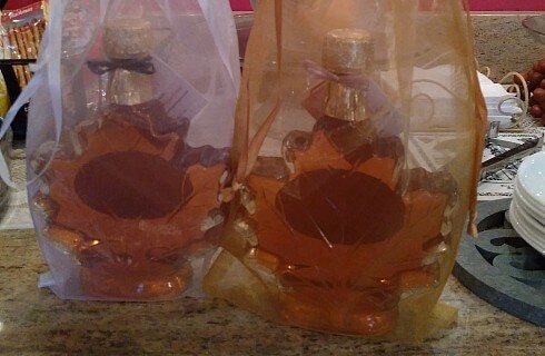 Two glass bottles of syrup in the shape of maple leaves inside fabric gift bags.