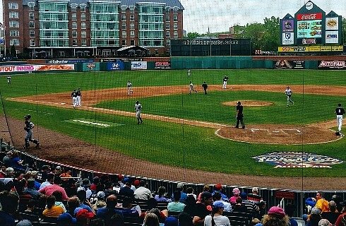 View from behind home plate of a baseball diamond with a crowd watching and apartment buildings in the background.