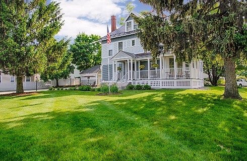 Two-story grey-blue home with white trim and large wrap around porch surrounded by lush green grass and trees.