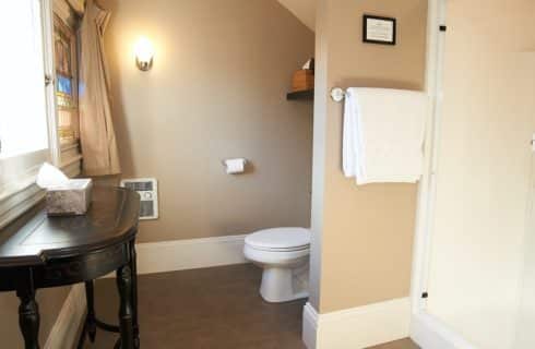 Bathroom with light beige walls, showing a toilet, shower, and small table under window with stained glass