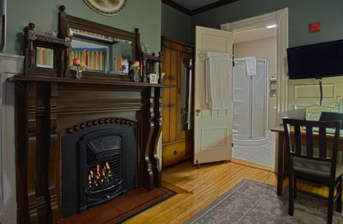 Beautiful dark brown wood mantle around a lit fireplace next to a sitting desk and an open door leading to a bathroom.