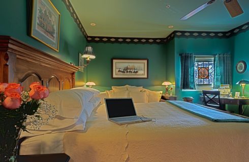 Bedroom with blue walls, bed with white linens and brown headboard and sitting desk in corner by a stained glass window.