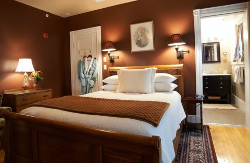 Bedroom with dark brown walls, queen bed with white linens, upholstered chair and view into the attached bathroom.