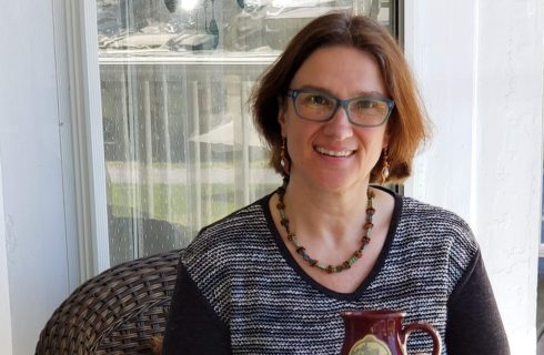 Woman with brown hair and blue glasses, wearing a black and white sweater, holding a mug while sitting outside on a wicker chair.