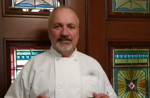 Man in a white chef's coat standing in front of large brown doors with colorful stained glass windows.