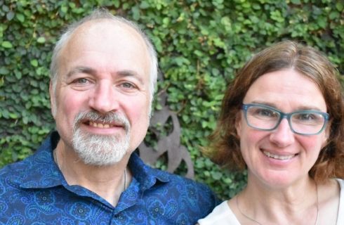 Man in a paisley blue collared shirt with grey hair and beard next to a brown-haired woman in blue glasses and white tank top.