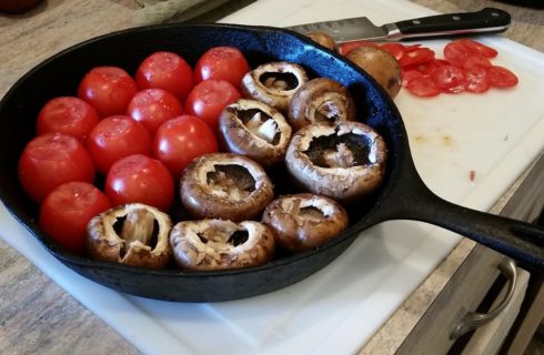 Black cast iron skillet on a table holding several whole red tomatoes and brown mushrooms.
