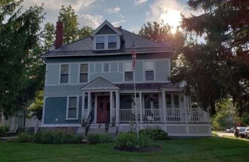 Front facade of a two-story home with light blue siding, white trim, wrap around porch and American flag in the lawn.