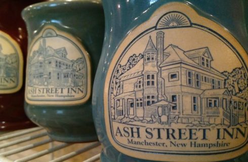 Three ceramic mugs; red, green and blue with an inn logo, sitting on a wire shelf.