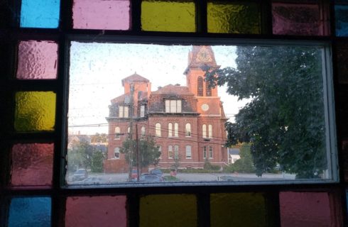 Blue, pink and yellow stained glass window looking across the street to a large red brick stately home.