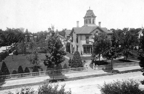 Black and white historical image showing stately home in a large lot with many trees and white fence