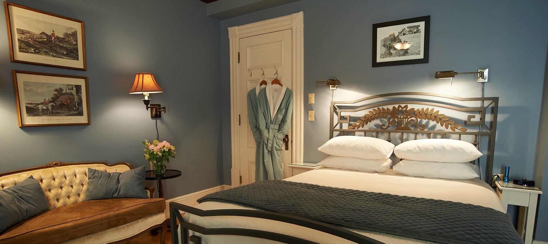 Bedroom with light blue walls, bed with white linens and iron frame, white tufted couch and two robes hanging on the door.