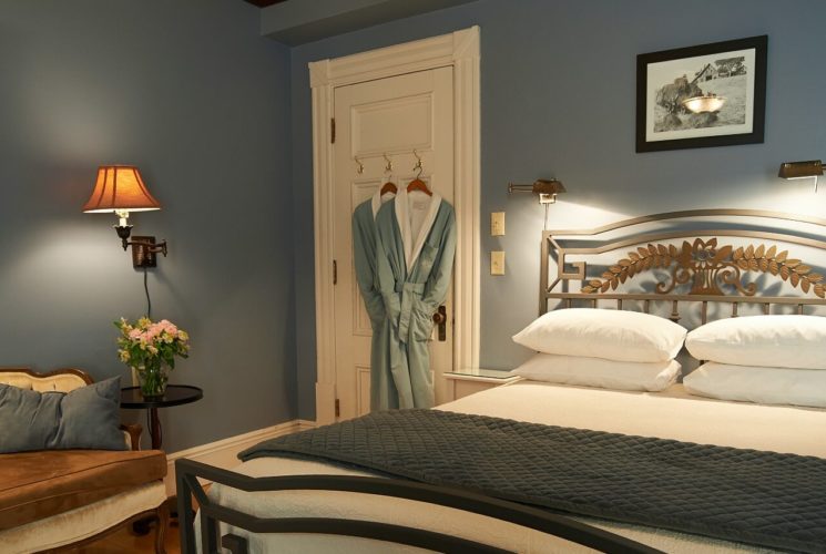 Bedroom with light blue walls, bed with white linens and iron frame, white tufted couch and two robes hanging on the door.