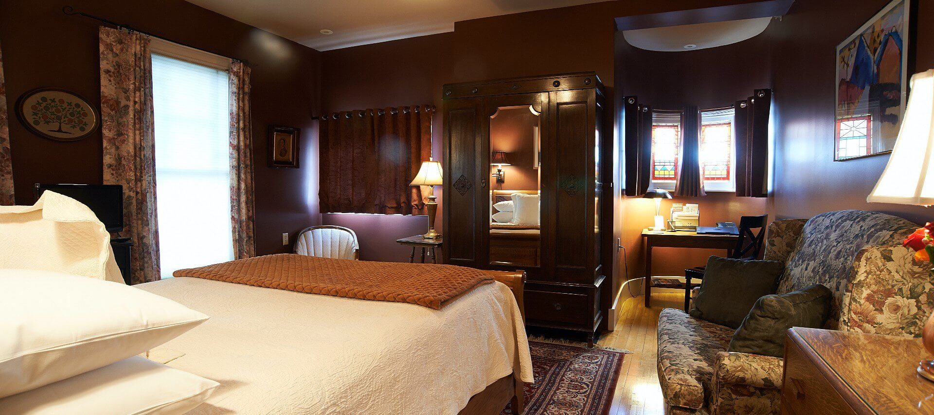 Large bedroom showing bed with white linens, dark brown walls, upholstered chair, sitting desk and large armoire.