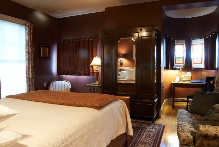 Large bedroom showing bed with white linens, dark brown walls, upholstered chair, sitting desk and large armoire.