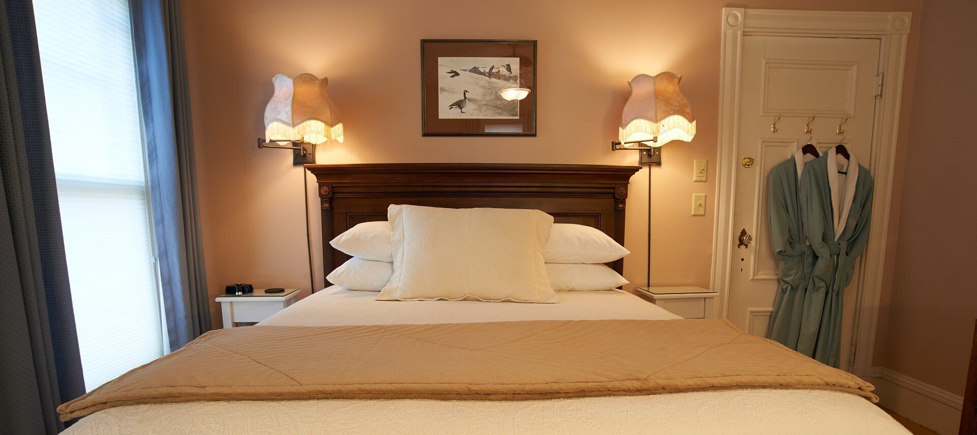 Bedroom with queen bed, white linens and tall dark brown headboard, bright window, two lamps lit and green robes hanging on the door.