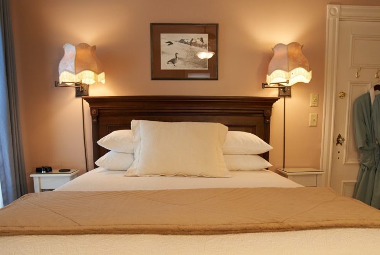 Bedroom with queen bed, white linens and tall dark brown headboard, bright window, two lamps lit and green robes hanging on the door.