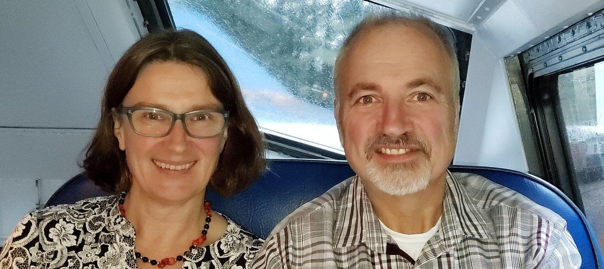 Woman with brown hair, glasses and black and white shirt sitting next to a man with grey hair and beard and black and white plaid shirt.