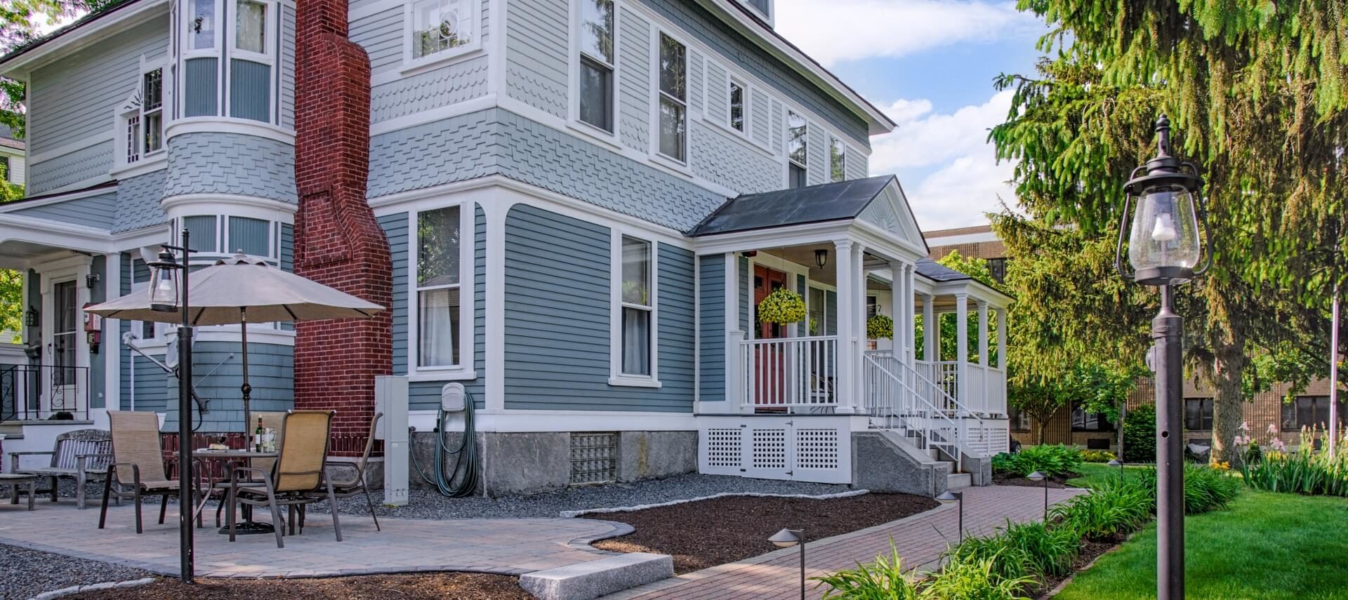 Outside of large home with slate blue siding, tall red brick fireplace, front porch and side patio sitting area.