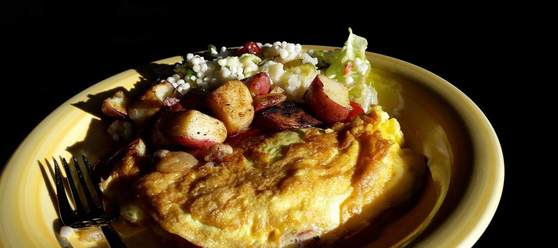 Yellow plate against a dark black background showing an omelette, diced redskin potatoes and silver fork.