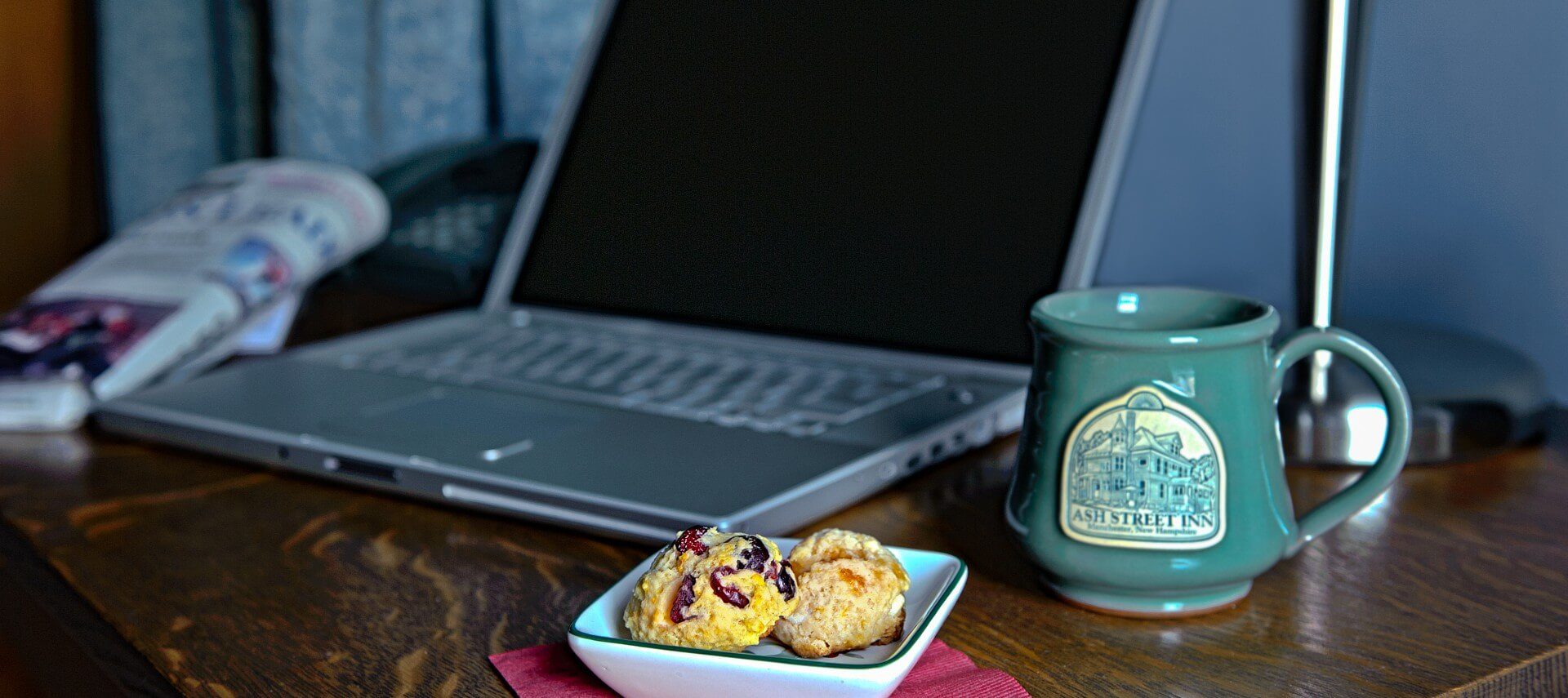 Brown desk with small plate of scones, green ceramic mug and an open silver laptop.