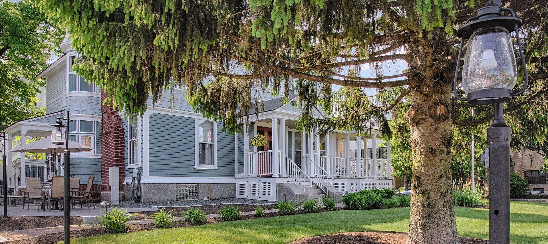 Beautiful home with grey-blue siding and white trim, with wrap around porch and patio sitting area next to grassy area and trees.