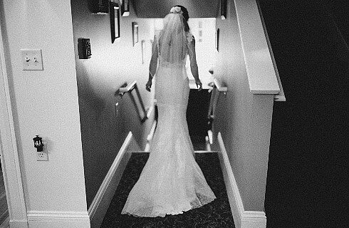 Backside of a bride wearing a white dress in veil standing in a hallway about to walk down a set of stairs.