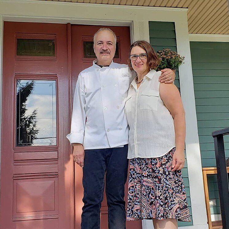 A woman in a white dressy tank top and floral skirt standing next to a man with jeans and white chef's coat at the front door of a home.