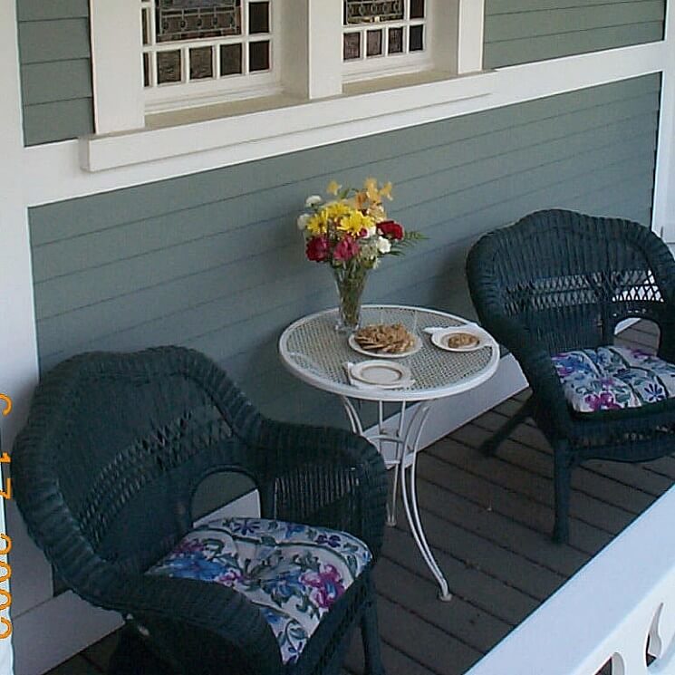 Two dark blue wicker chairs with floral cushions on a porch with a white side table holding flowers and snacks.