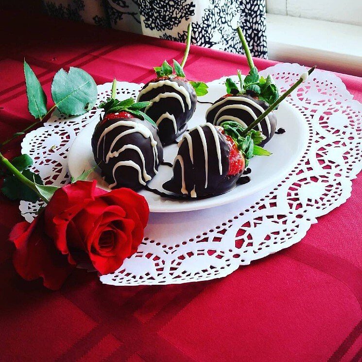 Table with red table cloth and lace doily holding a red plate with chocolate covered strawberries and one red rose.